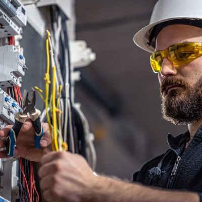 A male electrician works in a switchboard with an electrical connecting cable. Male electrician at the checkout counter on a blurred background of a switchboard.